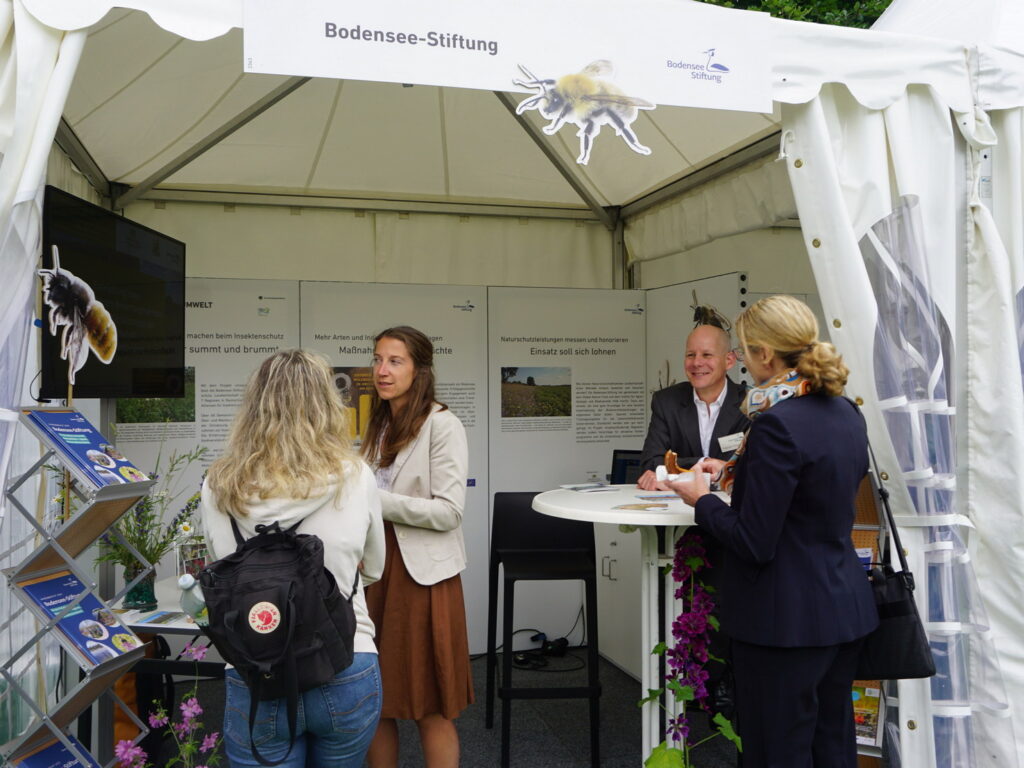 Auf dem Bild sind Projektleiterin Saskia Wolf und Geschäftsführer Patrick Trötschler von der Bodensee-Stiftung zu sehen. Sie stehen in einem Pavillon mit zwei weiteren Interessierten.