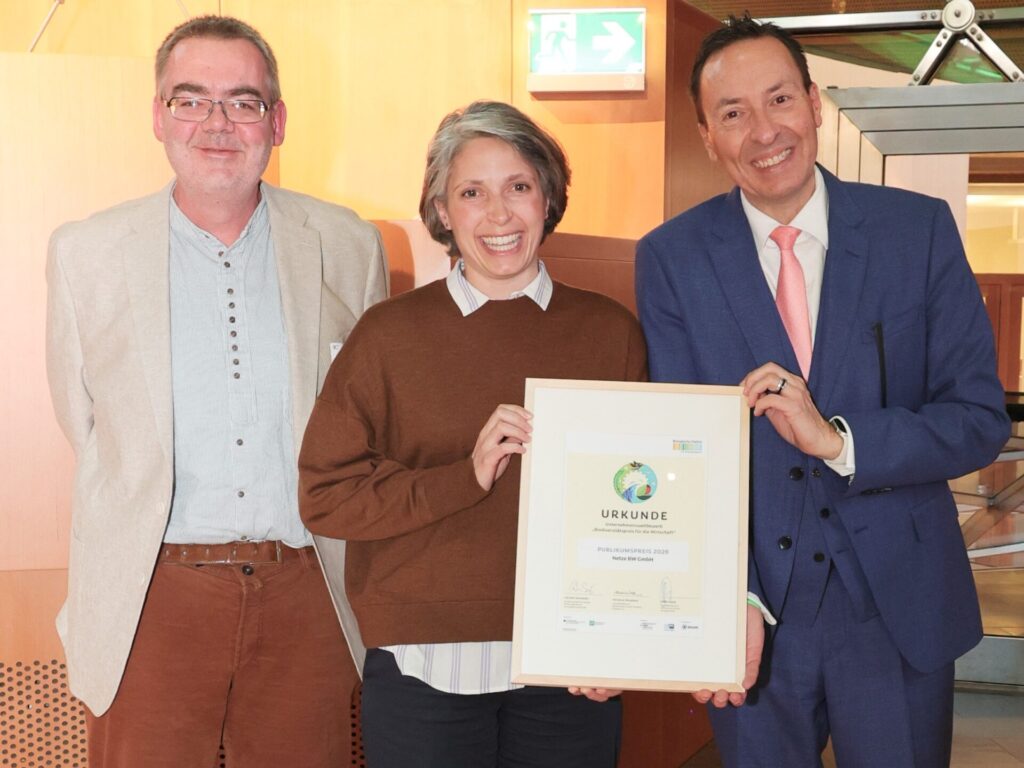 Eine Gruppe von drei Menschen schaut lachend in die Kamera. Die Frau in der Mitte hält eine Urkunde vor sich.