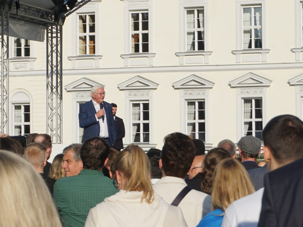 Zu sehen ist Herr Steinmeier auf einer Bühne. Er hält ein Mikrofon in der Hand. Im Vordergrund sind Menschen, die ihm zuhören.