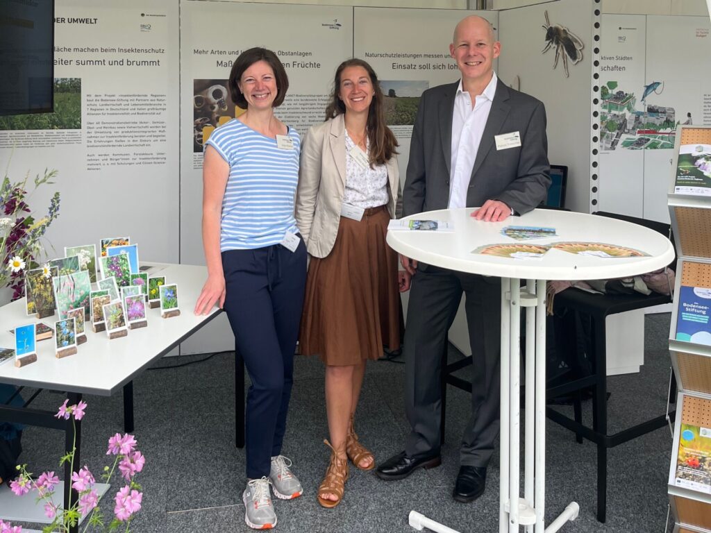 Auf dem Bild sind Anja Wischer (Kommunikation und Öffentlichkeitsarbeit), Saskia Wolf (Projektleiterin) und Patrick Trötschler (Geschäftsführer und Programmleiter Landwirtschaft und Lebensmittel) zu sehen. Sie stehen vor Plakaten und Bannern. Links ist ein Tisch mit kleinen aufgestellten Blumen Bildern. Rechts ist ein Stehtisch aufgebaut.
