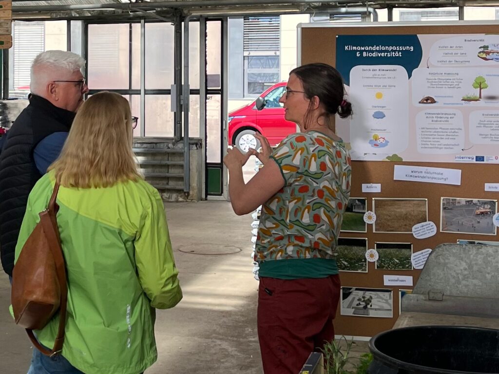 In der rechten Bildhälfte steht eine Frau vor einer Stellwand mit Poster. Sie unterhält sich mit zwei Menschen, die links stehen und auf die Stellwand schauen. 