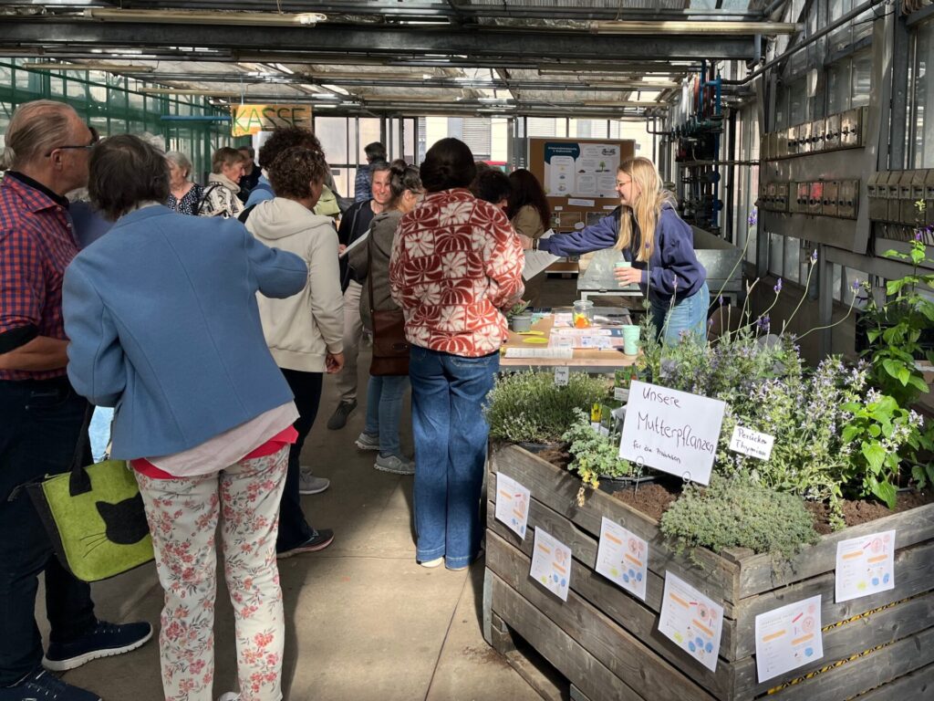 Menschen drängen sich in einem Gewächshaus vor einem Informationsstand mit Kräutern.