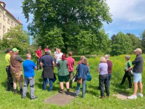 Zu sehen ist eine Gruppe vor einem großen grünen Baum auf einer Wiese.