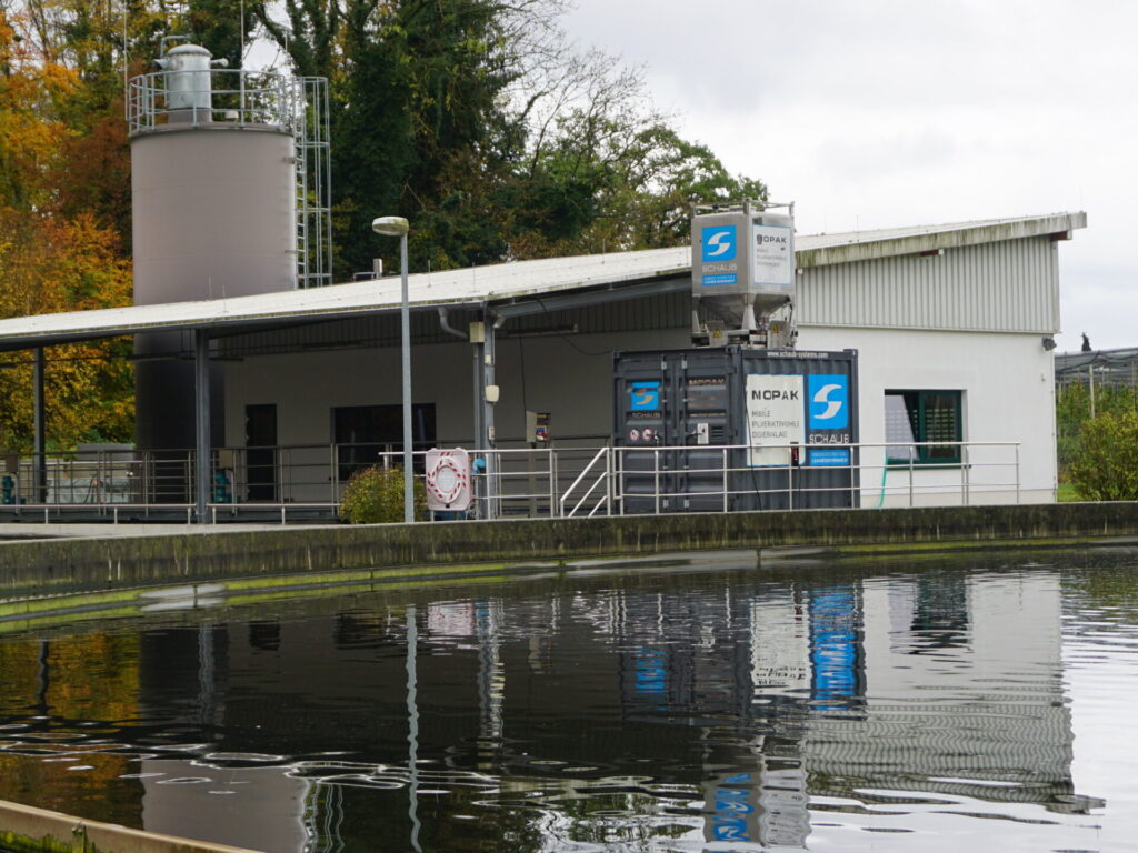 Man sieht ein rundes Becken. Dahinter ist ein Container, der vor dem Gebäude der Kläranlage steht.
