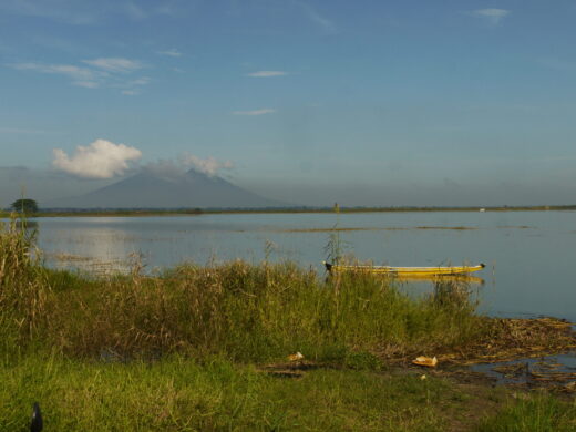 Ein See wird von der Sonne angescheint. Im Hintergrund sieht man einen Berg, der teilweise durch Wolken verdeckt wird. Auf dem See schwimmt ein Kanu.