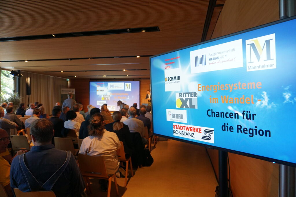 Vollbesetzter Veranstaltungssaal mit Blick auf eine Leinwand und einen Monitor mit dem Titel „Energiesysteme im Wandel – Chancen für die Region" sowie den Logos der Sponsoren.