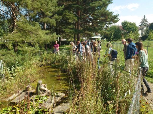 Mehrere Personen stehen entlang eines naturnahen Teiches in einem gepflegten Garten und betrachten die Wasserpflanzen und das Ufergrün. Im Hintergrund sind große Nadelbäume und Wohngebäude zu sehen.