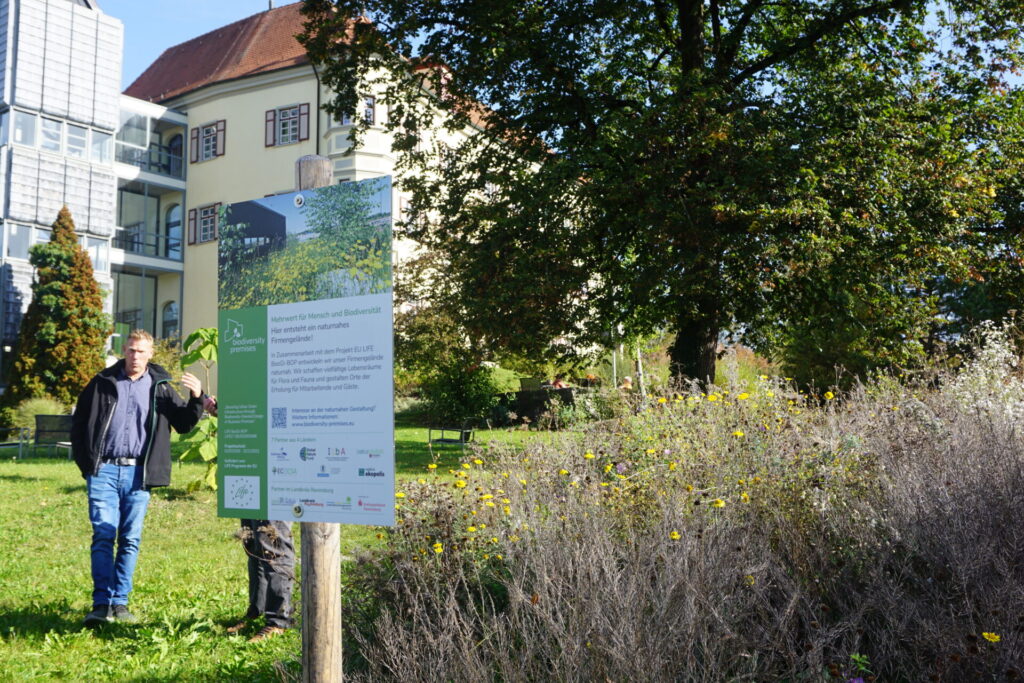 Im Vordergrund ist ein Schild. Dahinter sieht man ein Gebäude, vor dem ein großer Baum steht.