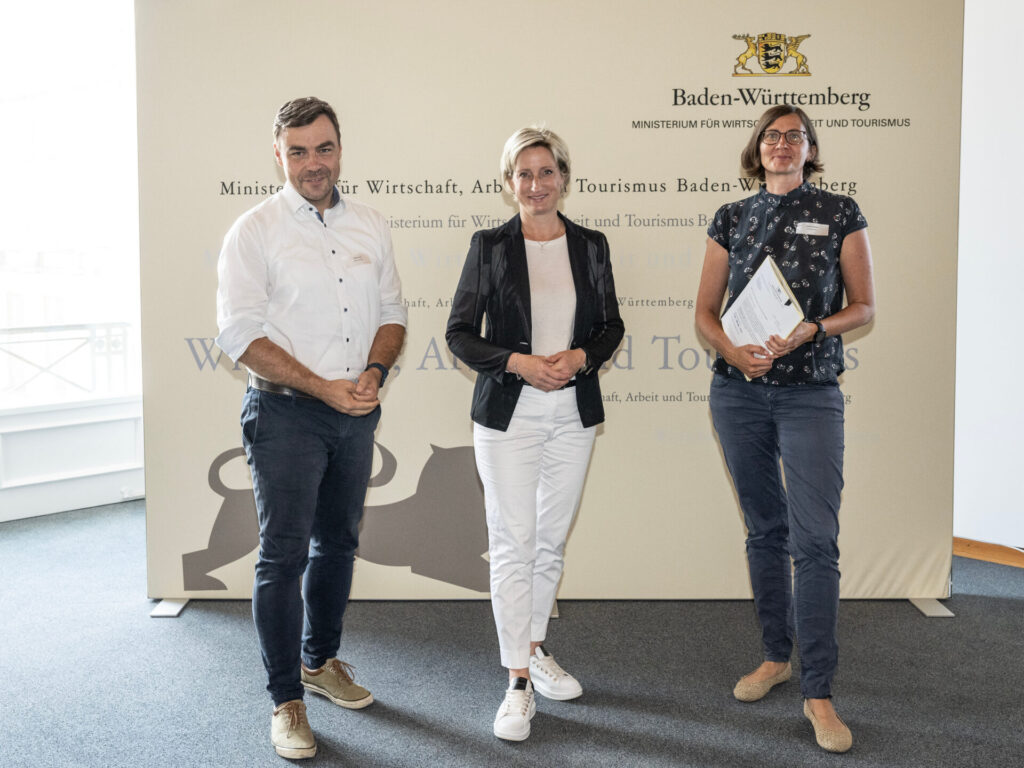 Drei Personen stehen vor einem großen Banner mit dem Logo des Ministeriums für Wirtschaft, Arbeit und Tourismus.