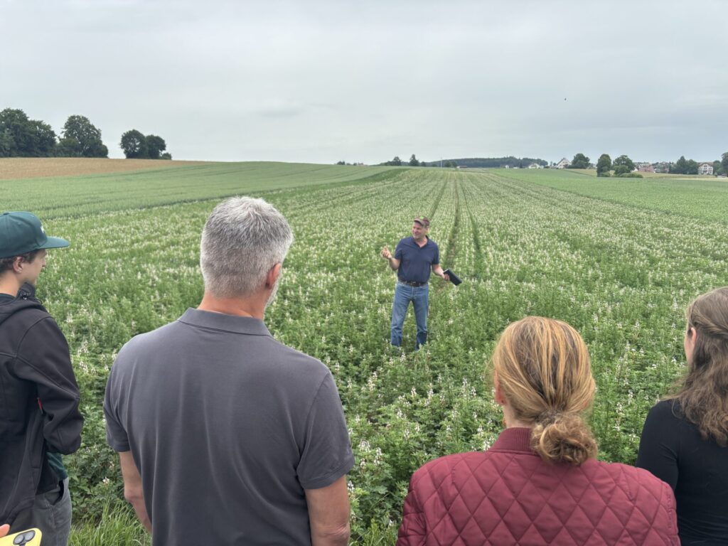 Eine Gruppe von Menschen ist von hinten zu sehen. Die Menschen stehen auf einem grünen Acker und blicken auf einen Landwirt, der vor ihnen steht und zu ihnen spricht.