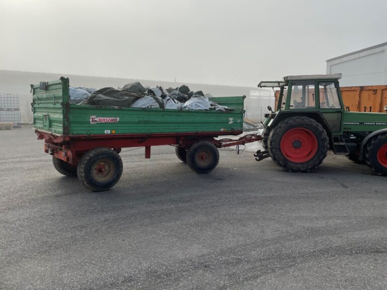 Ein Traktor steht mit einem Hänger auf einem Hof. Er hat gebrauchte Plastikfolie geladen.