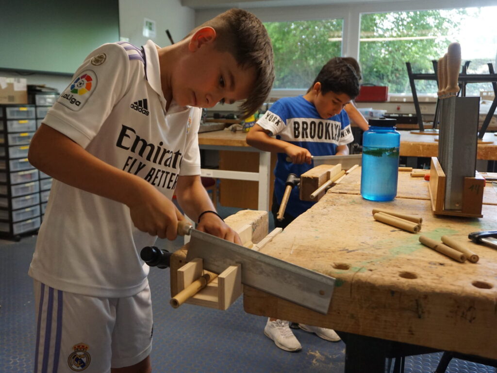 Ein Junge sägt ein Holzstab. Hinter ihm steht ein anderer Schüler.