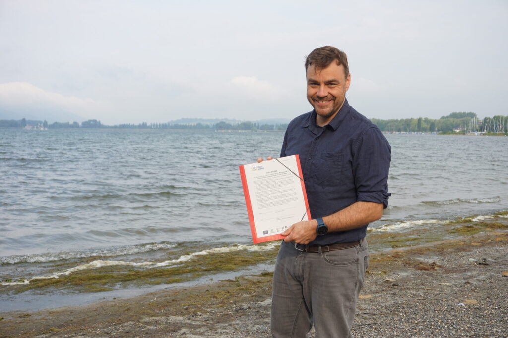 Dimitri Vedel steht vor dem Bodensee und hat das Seenpapier in der Hand.