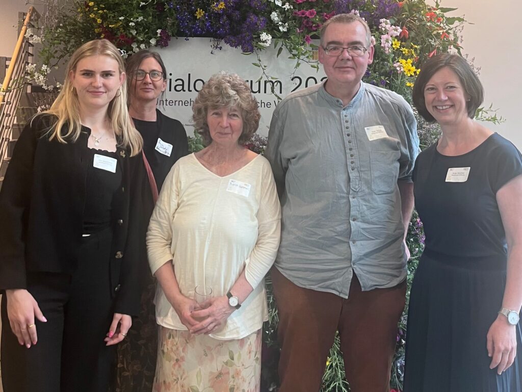 Eine Gruppe mit vier Frauen und einem Mann stehen vor einem mit Blumen umrankten Schild "Dialogforum 2026"