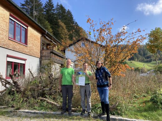 Drei Personen stehen vor Bäumen und Gräser. Die Frau in der Mitte hält ein Zertifikat in der Hand. Links im Bild sieht man eine Hausfassade.