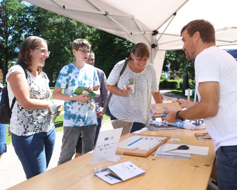 Unter einem Pavillon stehen Menschen. Einer davon ist hinter einem Tisch und erzählt den drei Personen vor dem Stand wichtige Infos rund um das Projekt.
