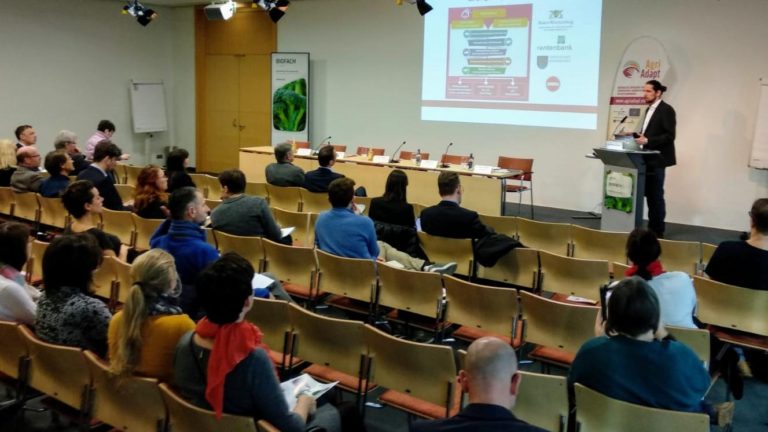Vortrag auf der Biofach-Messe: Ein Referent präsentiert am Pult vor etwa 40 Zuhörenden das EU-Projekt LIFE AgriAdapt. Auf der Leinwand ist eine Projektfolie zu sehen, rechts steht ein AgriAdapt-Rollup-Banner.