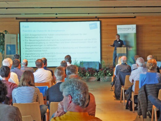 Vortrag bei der Fachveranstaltung zum Thema Bioenergie: Ein Referent spricht vor einem vollen Publikum, im Hintergrund eine Leinwand mit Präsentation und ein Roll-up mit Bioenergie-Logo.