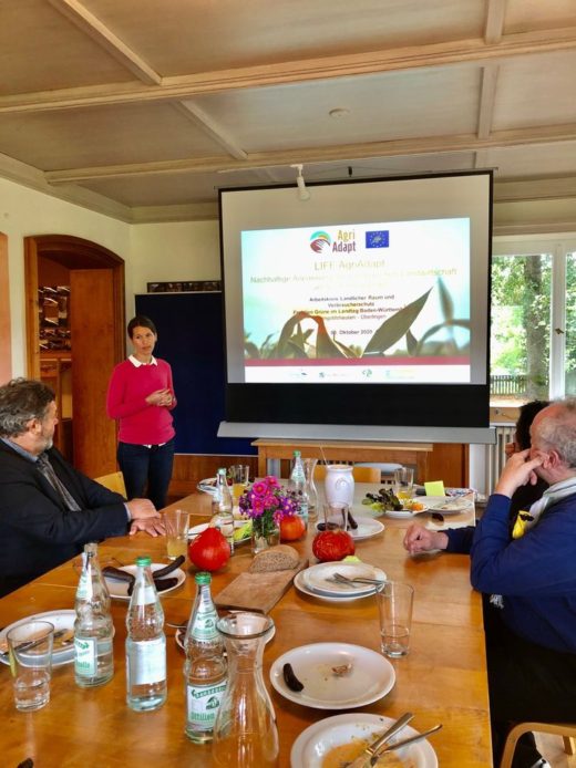 Eine Frau präsentiert das EU-LIFE-Projekt AgriAdapt vor einer kleinen Gruppe an einem Holztisch. Im Hintergrund zeigt eine Leinwand die Projektfolie „Nachhaltige Anpassung der europäischen Landwirtschaft".