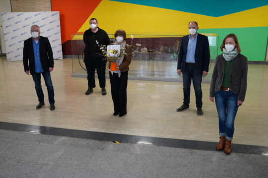 Fünf Personen mit Mund-Nasen-Masken stehen mit Abstand in einem Foyer vor einem farbenfrohen Wandbild – eine Person in der Mitte hält einen Blumenstrauß.