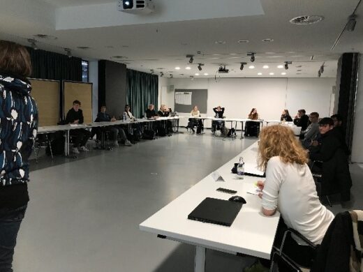 Zusehen ist ein Seminarraum. Viele Personen sitzen U-förmig ausgerichteten Tischen.