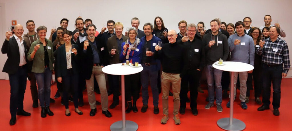 Gruppenbild auf einem roten Boden. Davor stehen zwei weiße Stehtische. Aller Personen haben ein Glas in der Hand zum anstoßen und heben dieses leicht nach oben.
