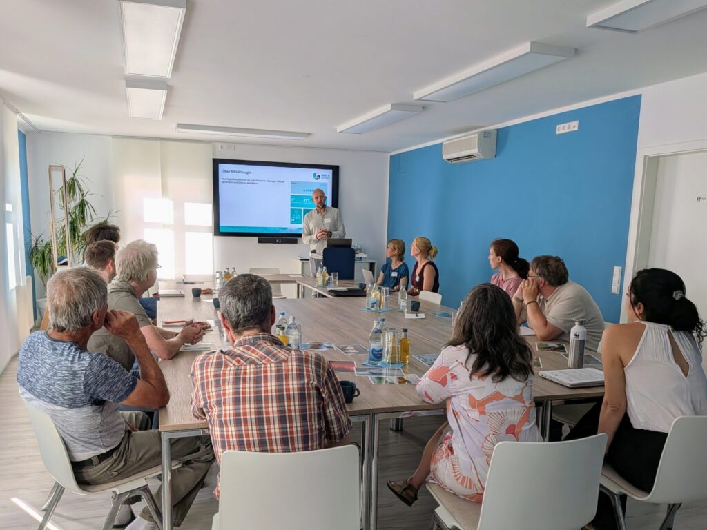 Eine Gruppe von 13 Menschen sitzt in einem Raum um einen Tisch und blickt auf einen Mann, der vor einem großen Bildschirm spricht. Auf dem Bildschirm ist die Folie mit der Überschrift "Über NBS4Drought" zu sehen.