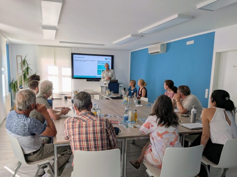 Eine Gruppe von 13 Menschen sitzt in einem Raum um einen Tisch und blickt auf einen Mann, der vor einem großen Bildschirm spricht. Auf dem Bildschirm ist die Folie mit der Überschrift "Über NBS4Drought" zu sehen.