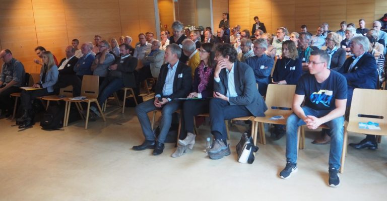 Gut gefüllter Veranstaltungssaal mit zahlreichen Teilnehmenden, die einer Präsentation folgen und sich untereinander austauschen.