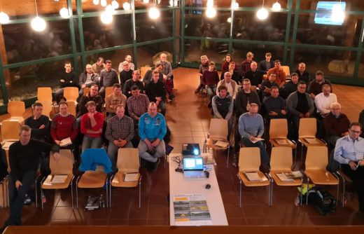 Vogelperspektive auf eine Gruppe von etwa 30 Personen, die in Stuhlreihen in einem Veranstaltungsraum sitzen. Im Vordergrund ein Tisch mit Laptop, Projektor und Informationsmaterialien.