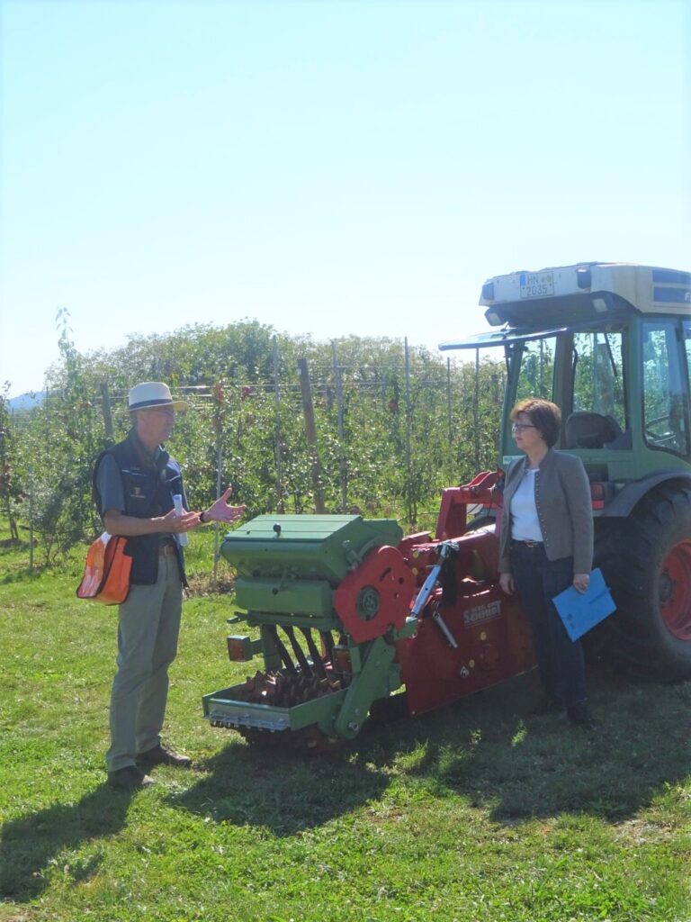Referent mit Strohhut erklärt einer Frau mit Mappe eine rote Sämaschine, die an einem grünen Traktor angehängt ist, im Hintergrund eine Obstplantage.