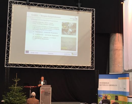 Ein Referent steht an einem Rednerpult und präsentiert auf einer Konferenz. Im Hintergrund ist eine Leinwand mit einer Folie zum Thema „ISABEL – Biogas-Kooperationen" zu sehen.