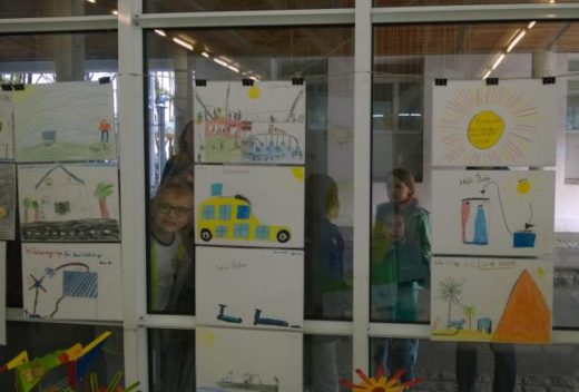 Kinder schauen durch eine Glasscheibe, an der Kinderzeichnungen zum Thema erneuerbare Energien hängen – darunter Windräder, Solarhäuser und ein Elektroauto.