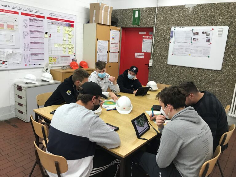 Sechs Jungs mit Maske sitzen an einem großen eckigen Tisch. Sie haben Tablets in der Hand.