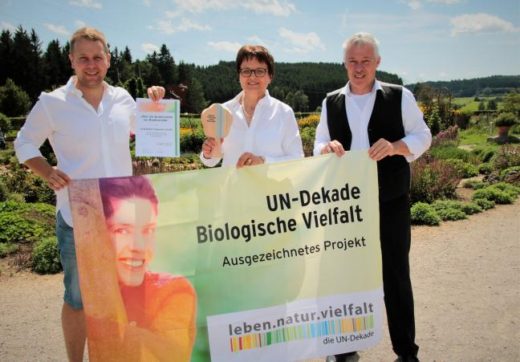 Drei Personen halten ein Banner in der Hand und lächeln in die Kamera.
