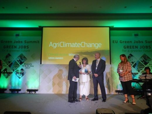 Preisverleihung beim EU Green Jobs Summit 2017 in Brüssel: Eine Frau erhält den Green Award für das Projekt „AgriClimateChange" von einem Moderator überreicht. Im Hintergrund eine grün beleuchtete Bühne mit der Projektbezeichnung auf der Leinwand.