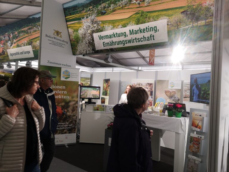 Messestand: Drei Besucherinnen und Besucher schauen sich den Stand an, eine Standbetreuerin steht dahinter. Links ein Roll-up zum Thema Insektenförderung in der Region.