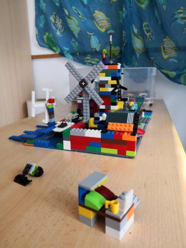 Aus bunten LEGO-Steinen gebaute Landschaft mit einer Windmühle und einem Windrad auf einer Grundplatte – ein Modell für Erneuerbare Energien, aufgestellt auf einem Holztisch.