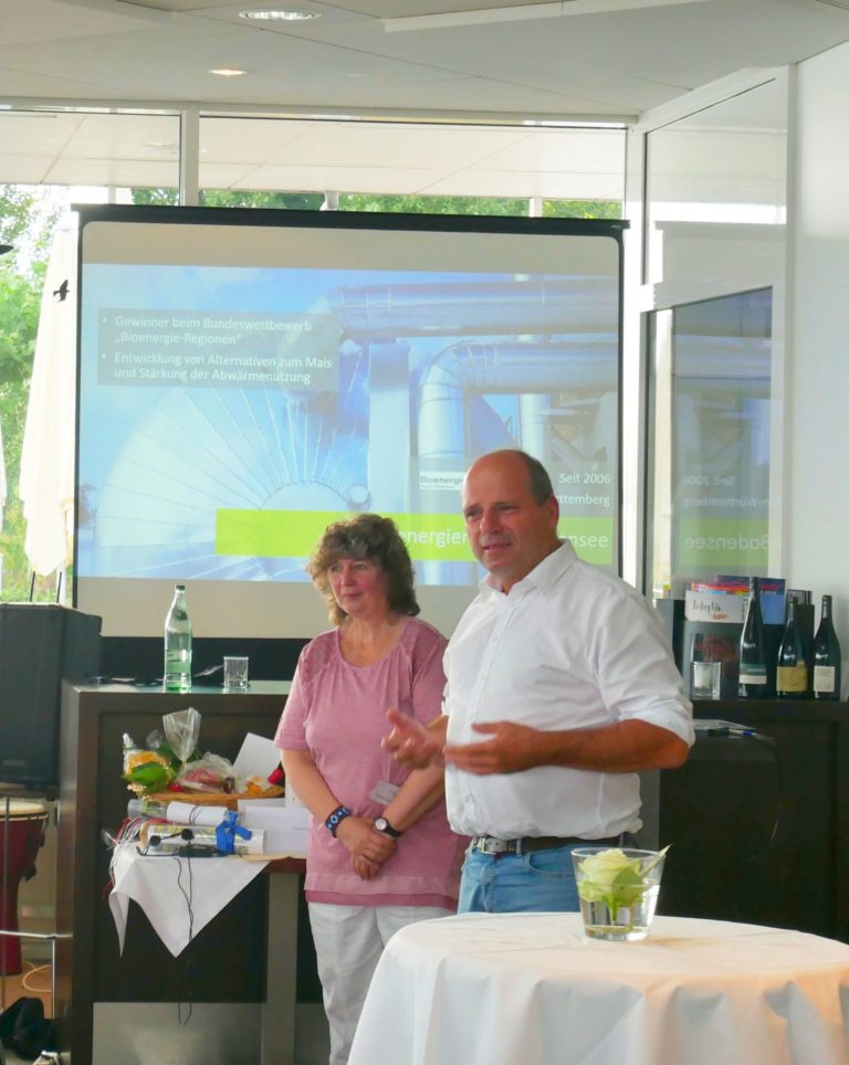 Zwei Personen stehen hinter einem Stehtisch. Hinter ihnen eine Leinwand mit einer PowerPoint Präsentation.