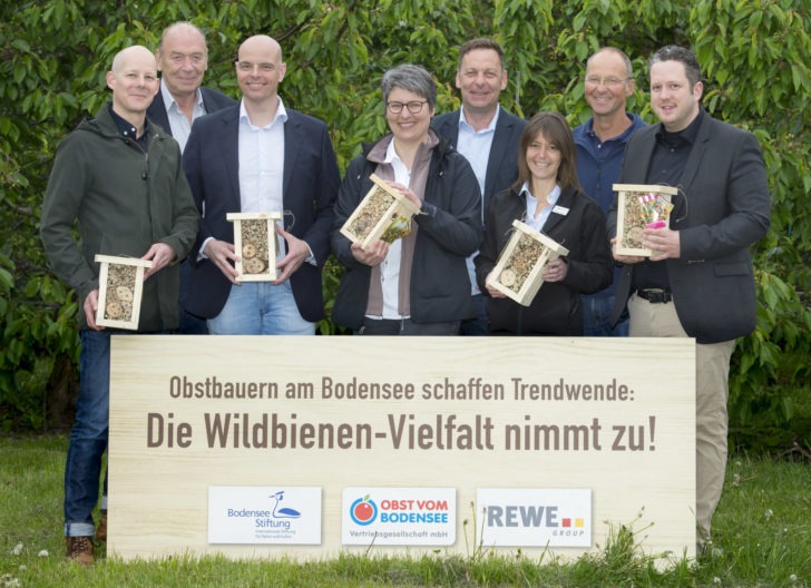 Acht Personen stehen vor einer Grünen Hecke und schauen in die Kamera. Sie halten jeweils Wildbienenhäuser in der Hand. Vor Ihnen steht ein Banner aus Holz mit der Aufschrift: Obstbauern am Bodensee schaffen Trendwende: Die Wildbienen-Vielfalt nimmt zu!