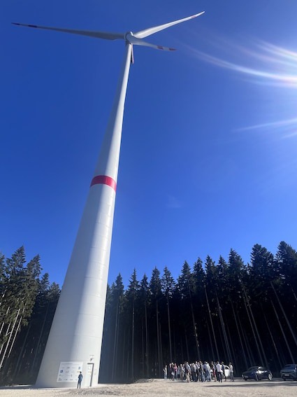 Hochformatige Aufnahme eines Windrads vor blauem Himmel. Am Fuß des Windrads steht eine Gruppe Menschen.