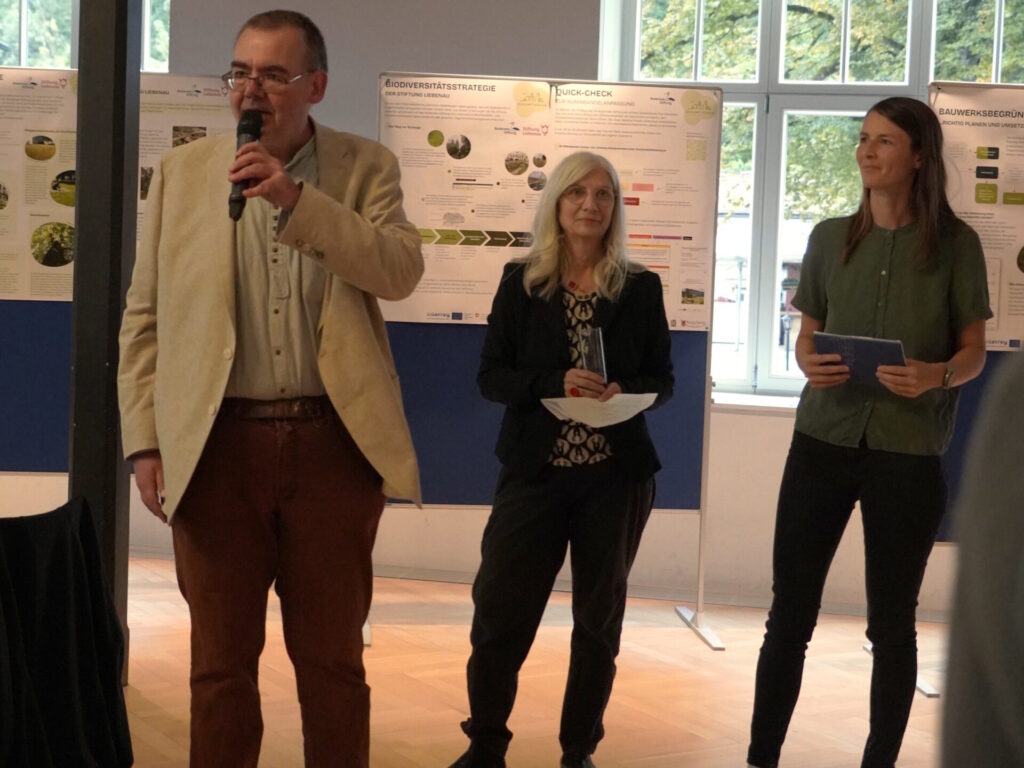 Drei Menschen stehen mit dem Rücken vor einer Posterwand und halten Notizen in ihren Händen. Der Mann links spricht in ein Mikrovon, neben ihm rechts stehen zwei Frauen.