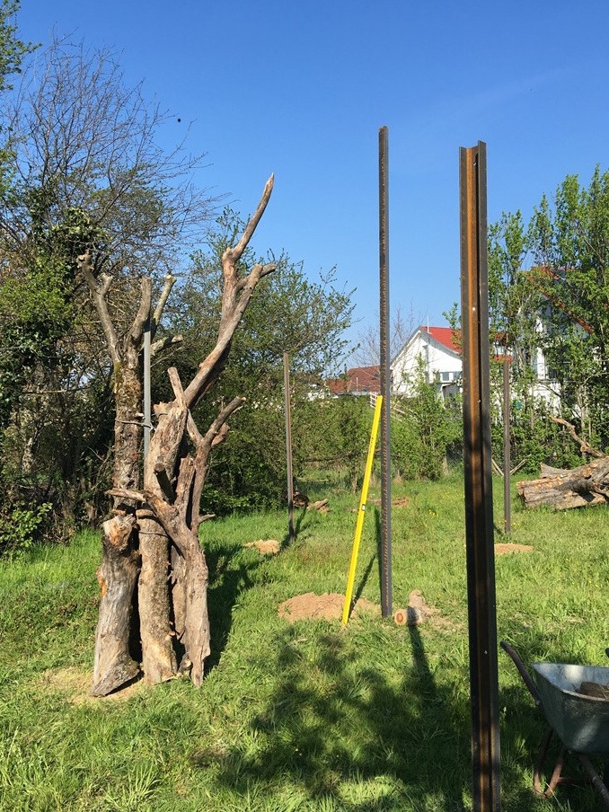 Blick in einen Garten. Rechts stehen Metallstäbe, links steht ein Bündel von Baumstämmen ohne Äste, die zusammengebunden sind.