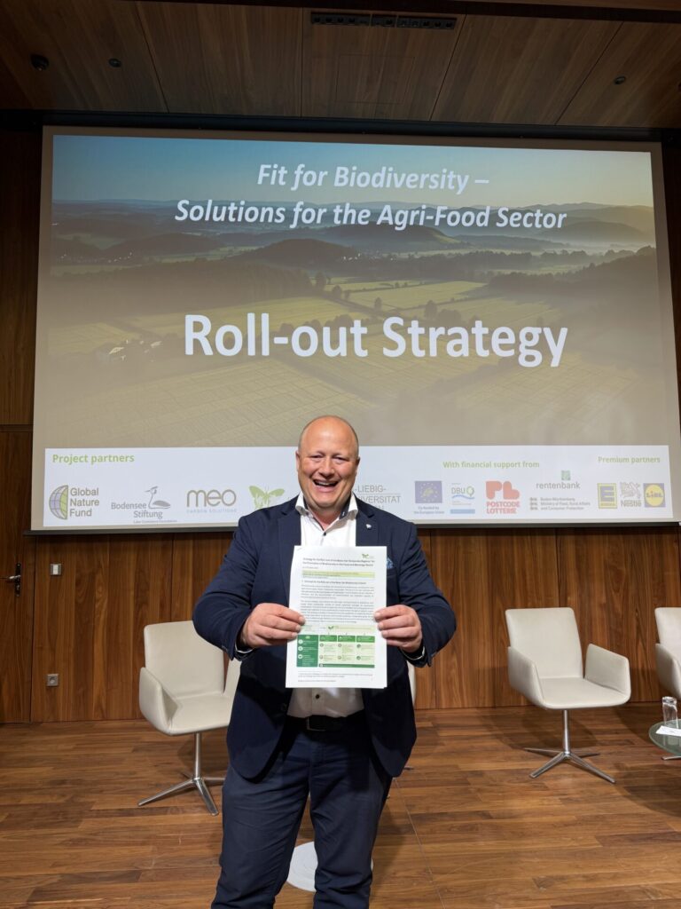 Ein Mann steht auf der Bühne eines Konferenzraums vor einer Leinwand, auf der steht: Roll-out-Strategie. Er schaut in die Kamera und hält ein Dokument vor sich in die Kamera.