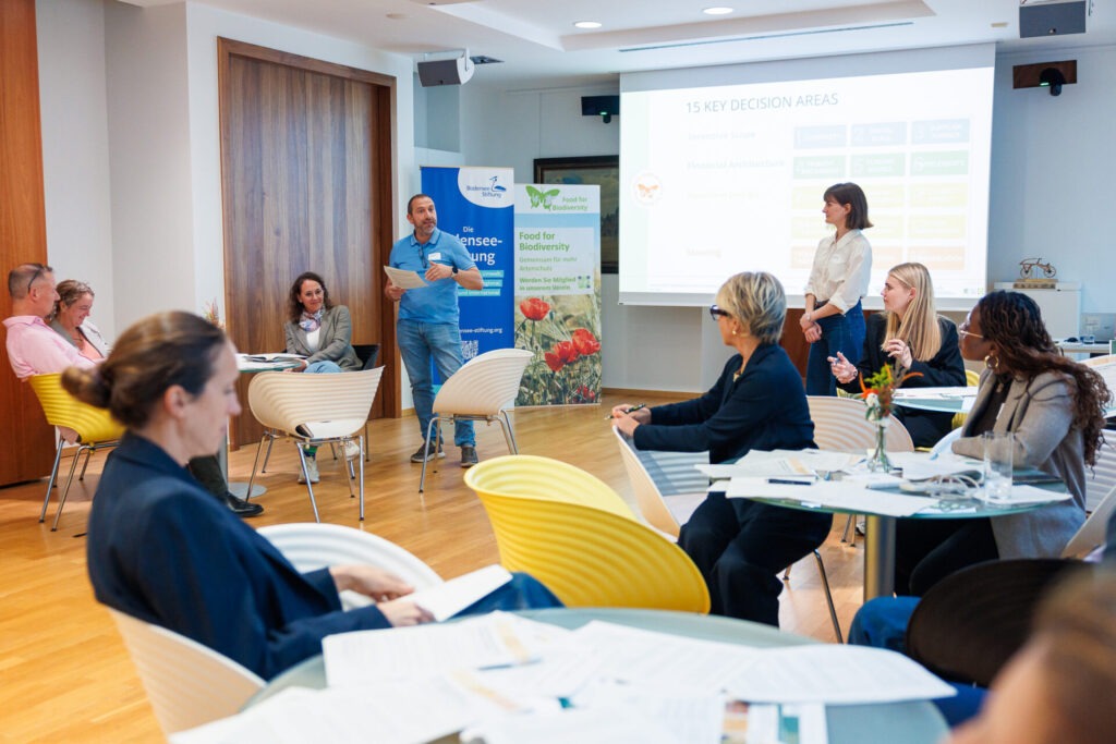 Workshopsszene in einem Konferenzsaal: Menschen sitzen an Kleintischen zusammen. Ein Mann steht in der linken Bildhälfte und spricht zu den Workshopteilnehmer*innen. Alle schauen zu ihm.