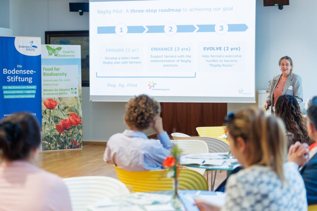 Eine Frau steht in enem Konferenzsaal vor einer Leinwand und spricht zum Publikum, das ihr zuhört.