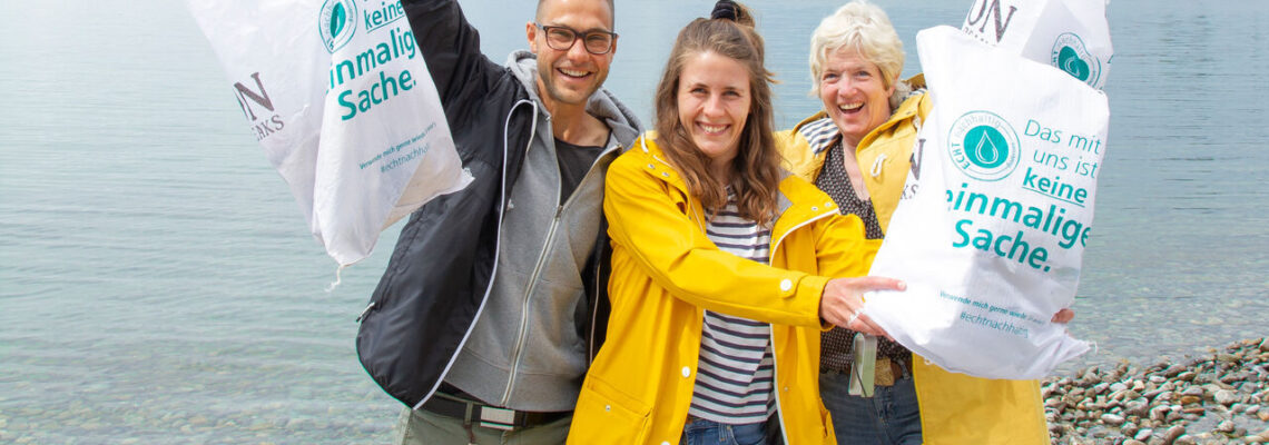 Ein Mann und zwei Frauen in Regenjacken stehen am Bodenseeufer und halten lachend Säcke hoch, die sie mit Müll gefüllt haben.