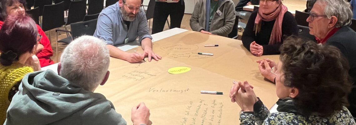 Eine Gruppe von Männern und Frauen sitzen um einen quadratischen Tisch. Sie diskutieren und machen Notizen auf einem Papier, mit dem der Tisch bespannt ist. Luisa Gedon steht hinter dem Tisch und verfolgt die Diskussion.