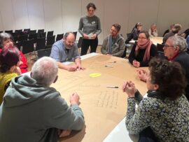 Eine Gruppe von Männern und Frauen sitzen um einen quadratischen Tisch. Sie diskutieren und machen Notizen auf einem Papier, mit dem der Tisch bespannt ist. Luisa Gedon steht hinter dem Tisch und verfolgt die Diskussion.