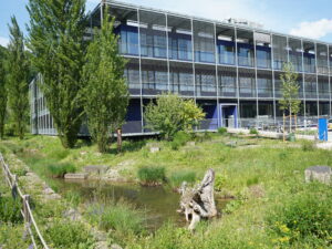 Blick auf ein Firmengebäude, das im Grünen steht. Vor dem Gebäude befinden sich Weiher, Bäume und Wiese